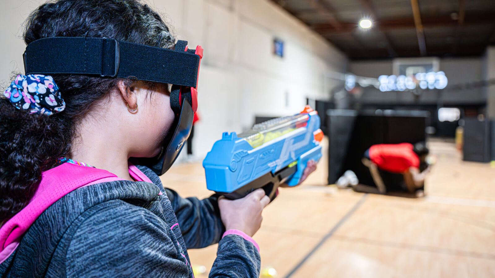 Child in protective goggles aiming a foam blaster during a Nerf battle on the court